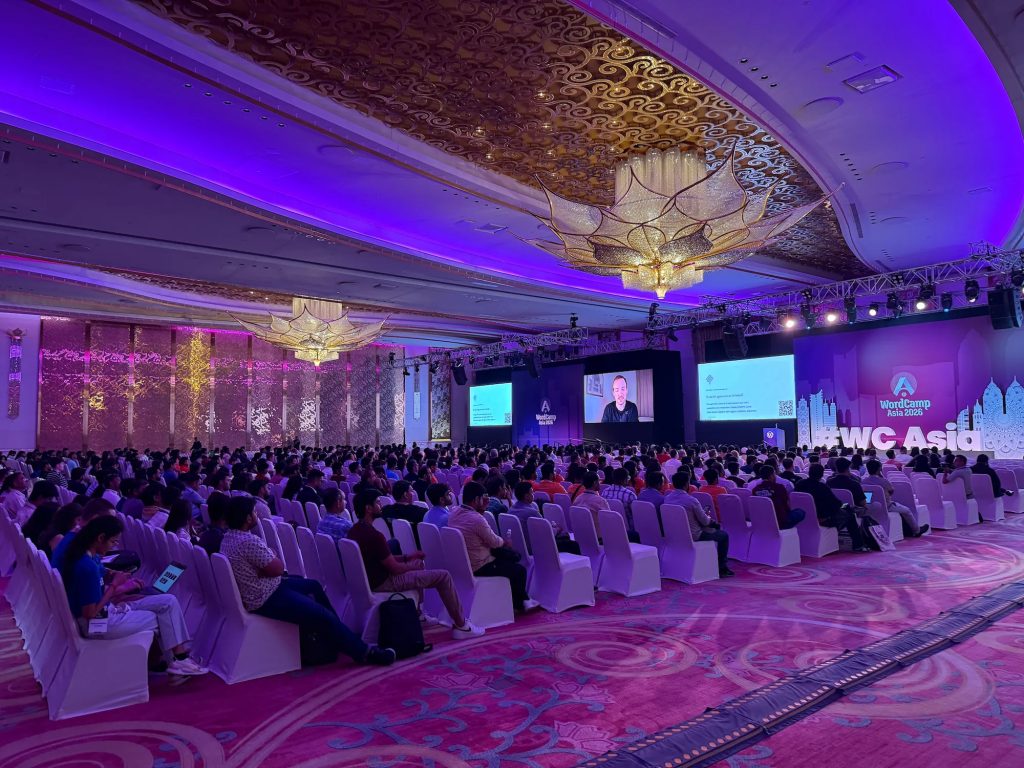 James L. apresenta remotamente em um telão para uma grande plateia em um salão iluminado de roxo no WordCamp Asia 2026